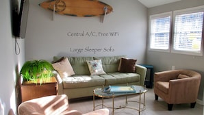 A living room with a large sleeper sofa, a glass coffee table, a pink armchair, a green plant, and a surfboard decoration on the wall. - LAVALLETTE New Panoramic Deck, Outdoor shower, and Kitchen INCLUDES BEACH BADGES (Lavallette)