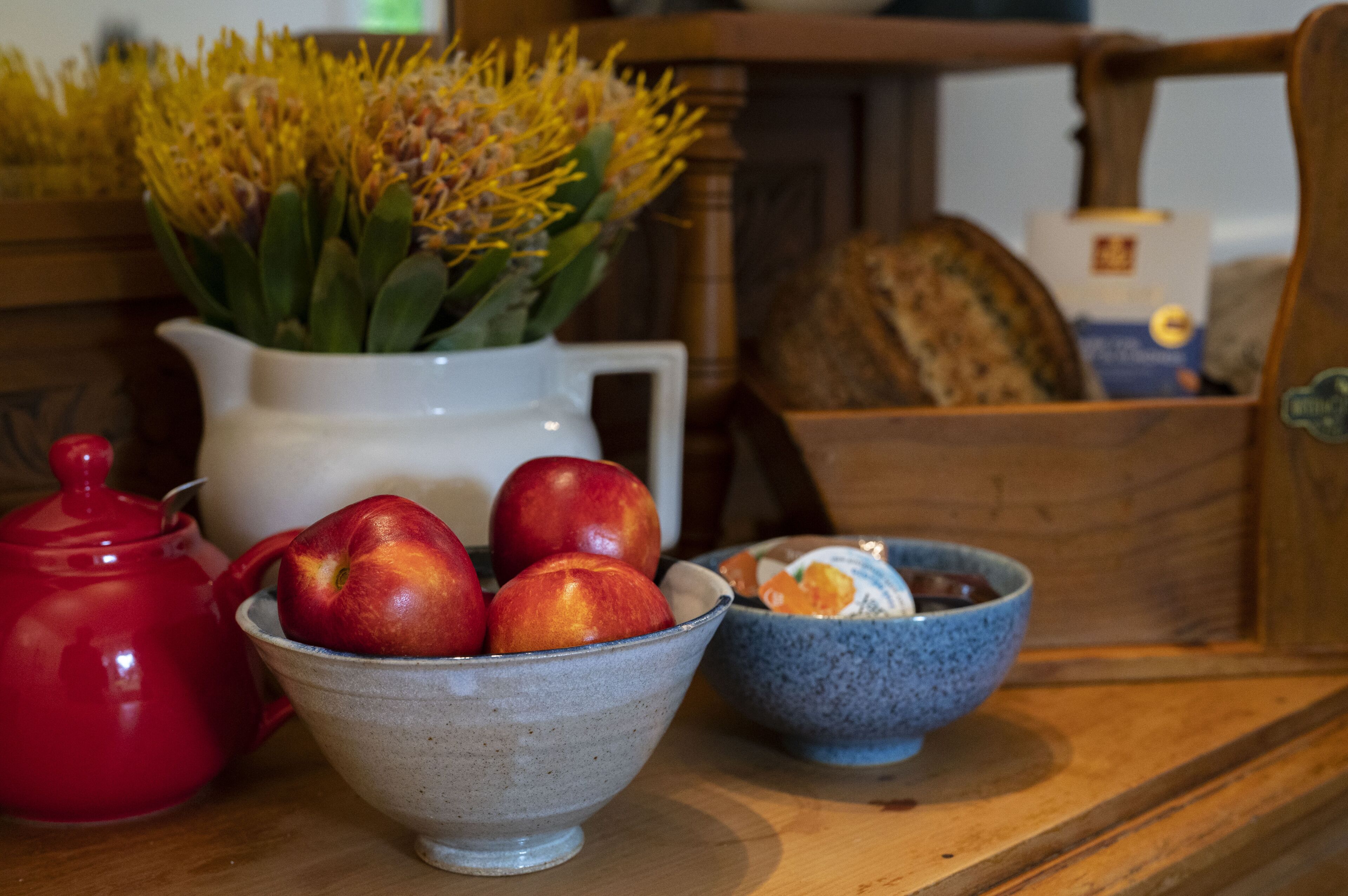 Une table en bois sur laquelle se trouvent un pichet blanc, une théière rouge, un bol de pommes, un bol de yaourt et un vase de fleurs.