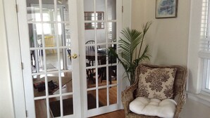 A room with a white door, a wicker chair with a cushion, a potted plant, and a framed picture on the wall.