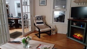 A living room with a wooden floor, a white sofa, a table with a vase of flowers, a fireplace, and a television on a stand.