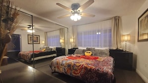 Un dormitorio con una cama grande, un ventilador de techo, un espejo y una ventana con cortinas.