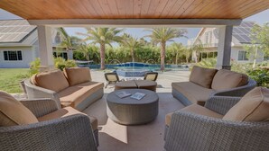 Un espacio de estar al aire libre con techo, amueblado con muebles de mimbre y una mesa redonda central, con vistas a una piscina y palmeras.