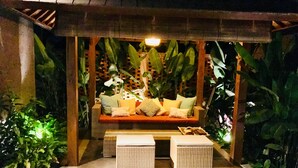A cozy outdoor seating area with a cushioned bench and two wicker ottomans, surrounded by lush tropical plants and a wooden lattice backdrop. - Welcome to Paradise. Quiet & Tranquil.  Pool & Garden  (Ubud)