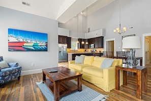 A spacious living room with a yellow sofa, a wooden coffee table, and a painting of boats on the wall.