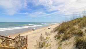 Une plage avec un escalier en bois menant au sable, une plage de sable bordée d’une clôture en bois, et l’océan en arrière-plan.