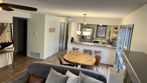 A modern living room with a gray sofa, a wooden dining table, and a kitchen area with a refrigerator and stove. - The Resting Place on Pine Cove at Mountain Harbor (Mt Ida)