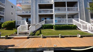 A multi-story residential building with balconies, a wooden deck, and a flag. - Cozy, inviting 1 bedroom condo with an outdoor pool and gorgeous canal-front view located uptown on the bayside just a few blocks from the beach! (Ocean City)