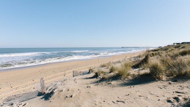 Ein Strand mit einer Holzumzäunung, Sanddünen und einem Pier in der Ferne.