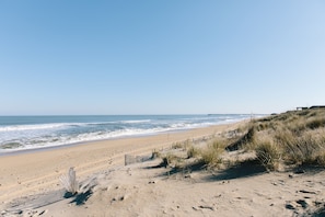 Uma praia com cerca de madeira, dunas de areia e um píer ao longe.