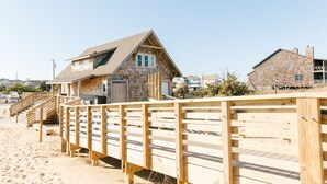 Ein hölzerner Steg führt zu einem Strandhaus mit geneigtem Dach und einer Steinfassade.