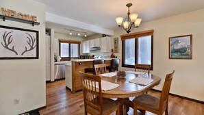 A dining area with a wooden table, chairs, and a chandelier. There is a kitchen in the background with white cabinets and a window.