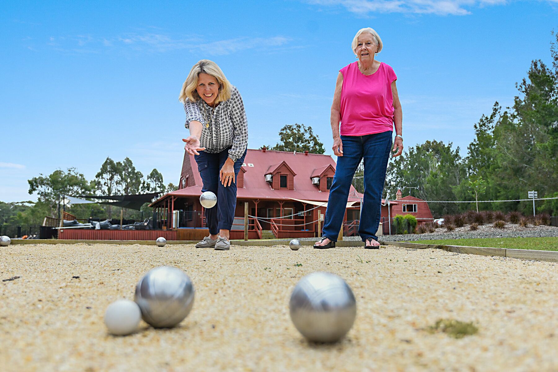 Dues persones jugant a croquet en un camp de grava amb un edifici amb teulada vermella al fons.