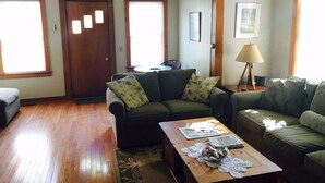 A living room with a wooden floor, two sofas with patterned cushions, a coffee table with magazines and a decorative bowl, and a lamp on a side table. - Locally owned downtown home:  Bar Harbors Crows' Nest (Bar Harbor)