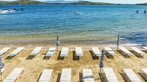 Ein Strand mit Reihen von Liegestühlen und Sonnenschirmen, einem klaren blauen Meer und einer bewaldeten Hügelkette im Hintergrund.