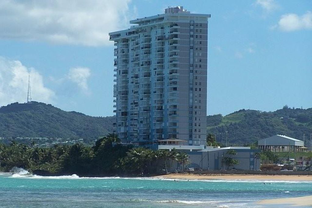 A tall, multi-story hotel with balconies, located near a beach with clear blue waters and a hilly backdrop.