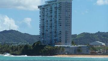 A tall, multi-story hotel with balconies, located near a beach with clear blue waters and a hilly backdrop.