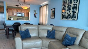 A living room with a beige sectional sofa, a dining table with chairs, a kitchen area with wooden cabinets, and a blue wall with decorative elements. - Luquillo, 18th Floor With Ocean Front  (Luquillo)