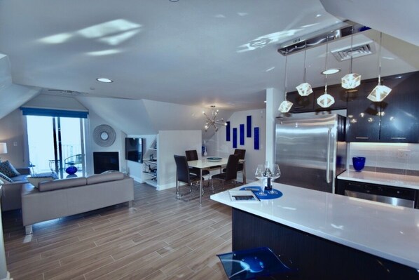 A modern kitchen with a central island, a dining area with a round table, and a living space with a sofa and a fireplace.