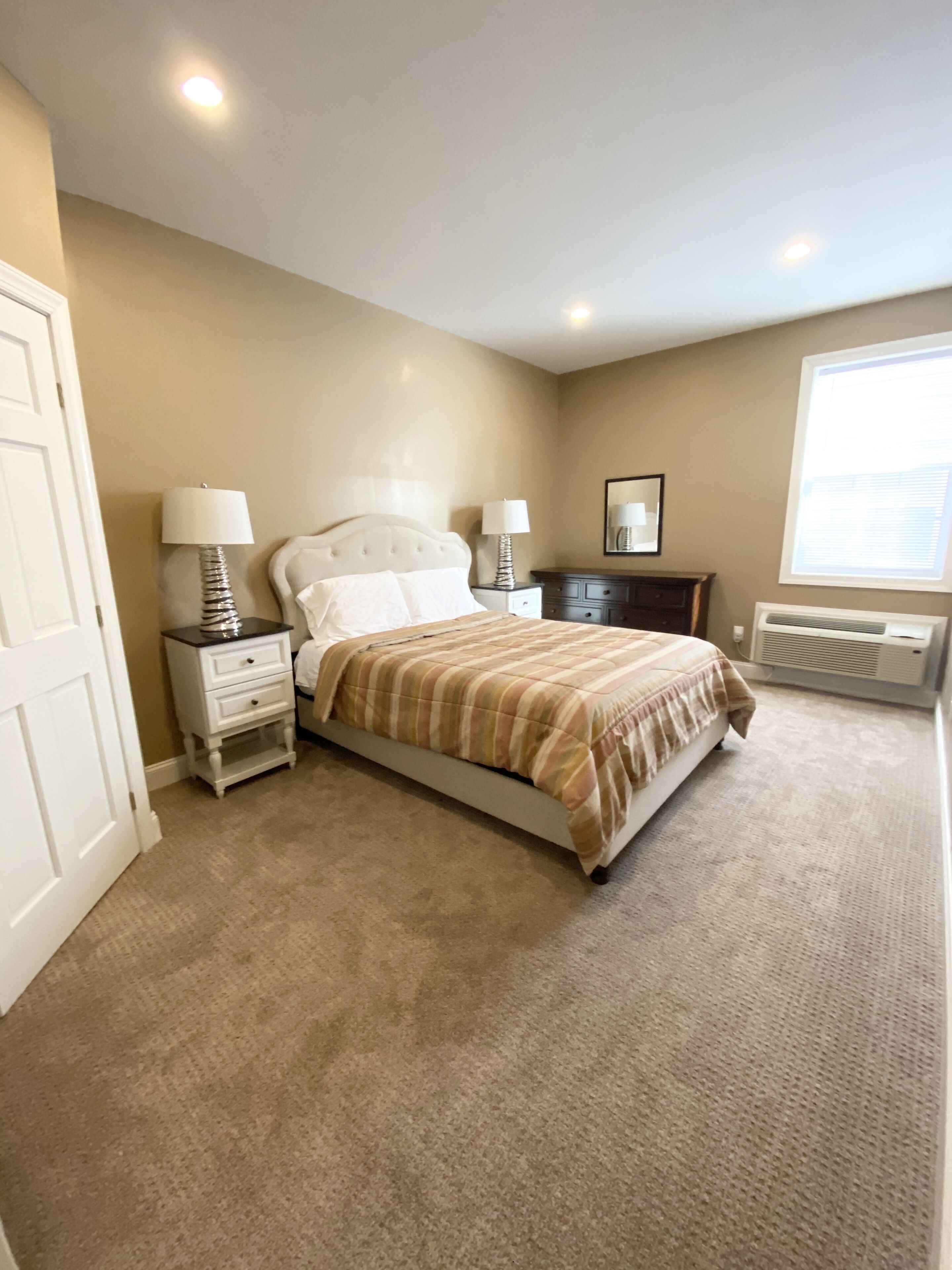 A bedroom with a bed, two bedside tables with lamps, a dresser, a mirror, and a window with blinds.