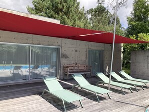Zona de piscina con sillones de descanso y un banco bajo un toldo rojo.