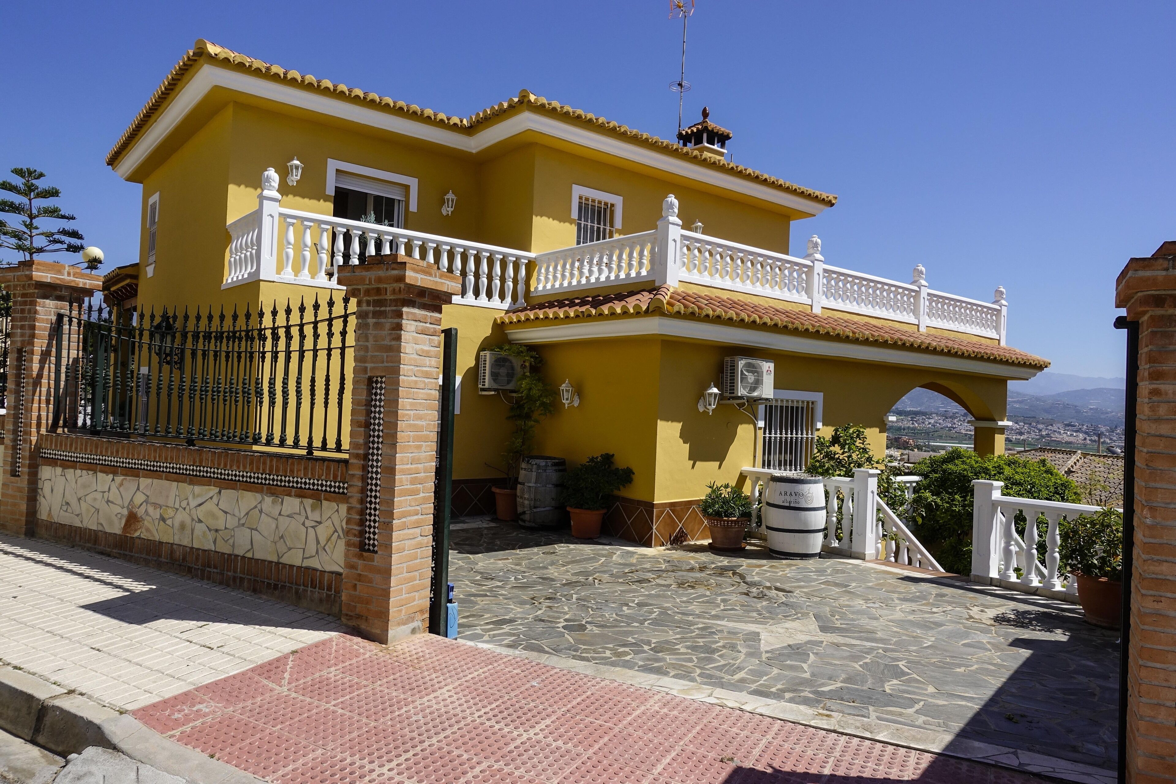 A yellow building with a balcony, a gated entrance, and a stone-paved driveway.