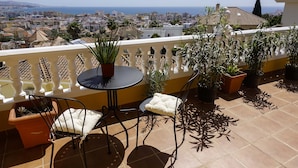 A terrace with a table and chairs, potted plants, and a view of a coastal cityscape.