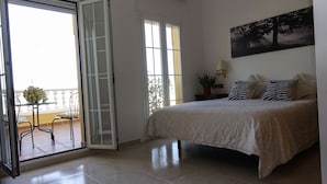 A bedroom with a bed, a sliding glass door leading to a balcony, a potted plant, and a painting on the wall.