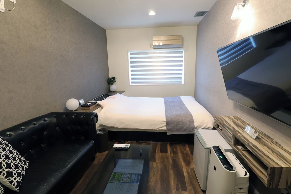 Une chambre d’hôtel de taille réduite, comprenant un lit, un canapé en cuir noir, une petite table, un téléviseur et une kitchenette compacte.