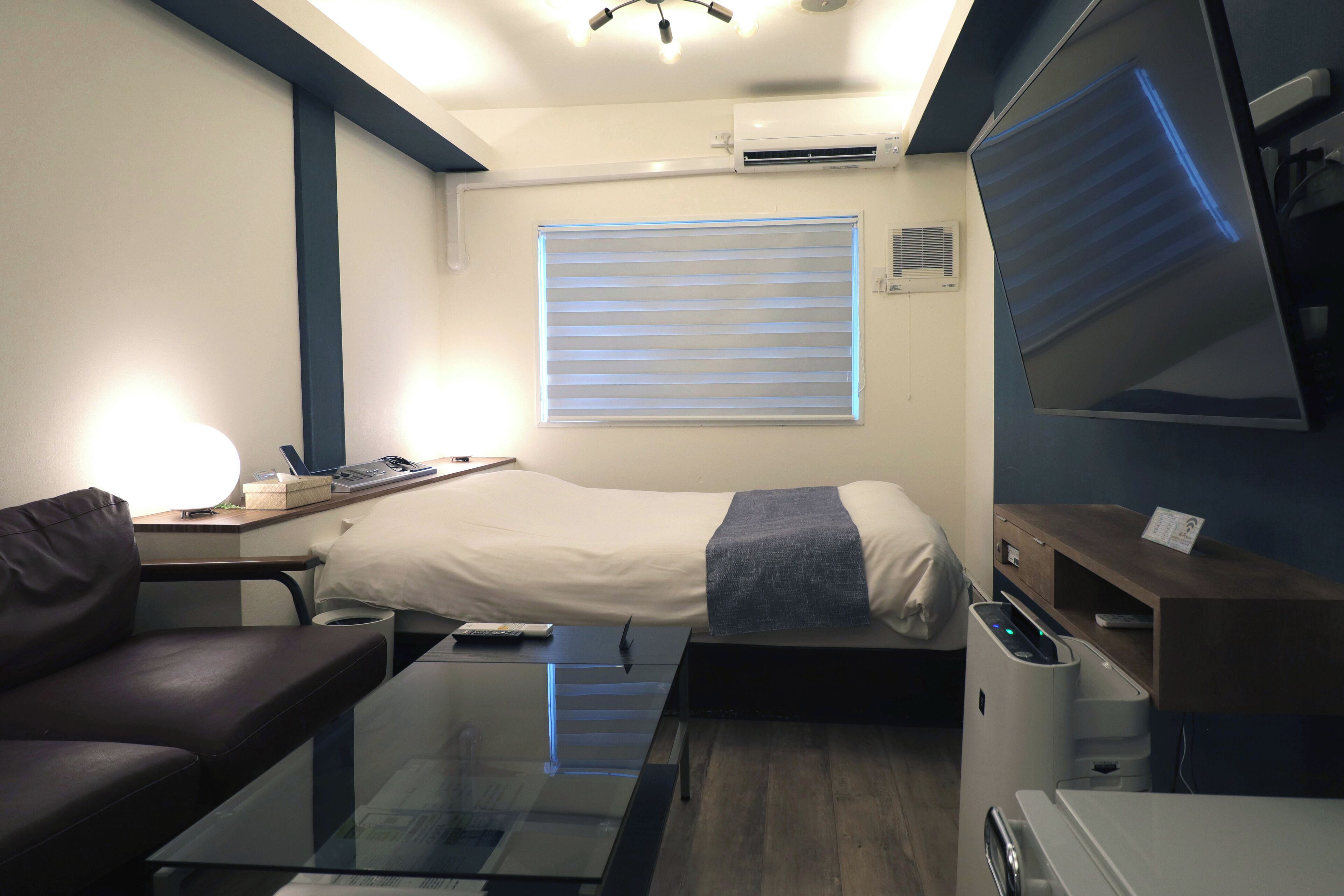 A compact hotel room with a bed, a desk with a lamp, a sofa, a TV, and a small table.