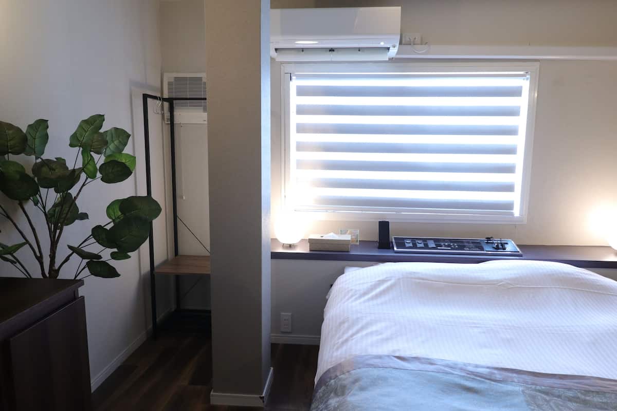Une chambre à coucher avec un lit, un bureau avec un ordinateur, une plante et une fenêtre avec des stores.