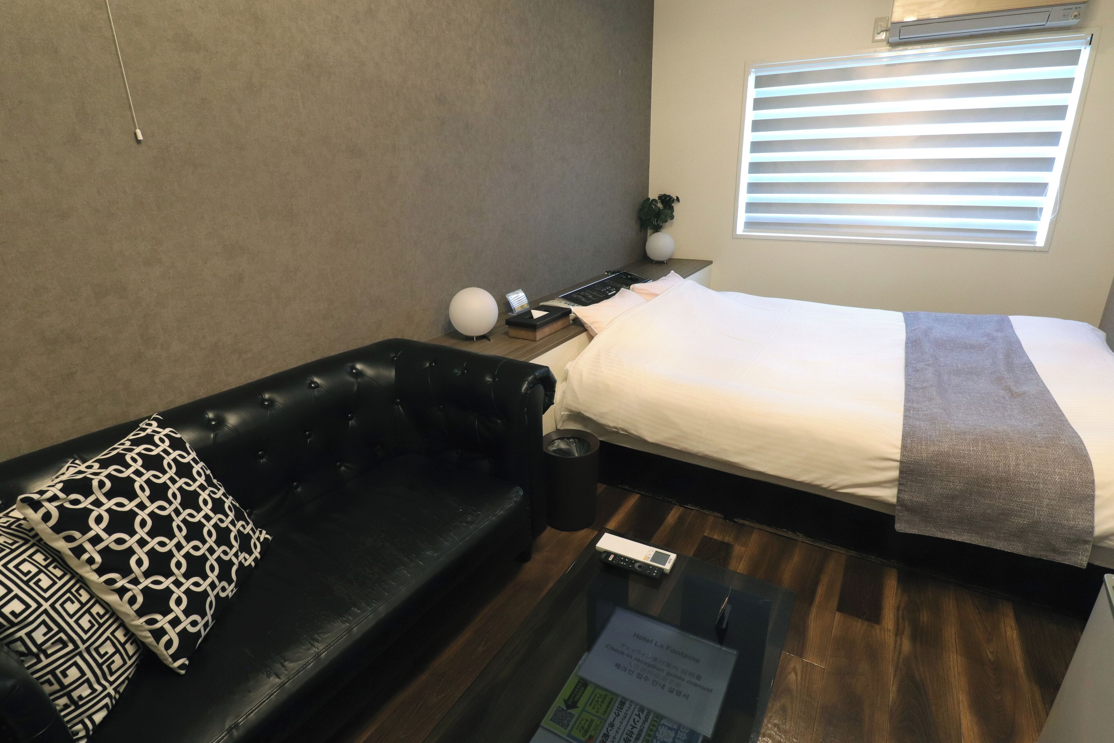 Ein Hotelzimmer mit Bett, einer schwarzen Ledersofa, einem kleinen Tisch und einem Fenster mit Jalousien.
