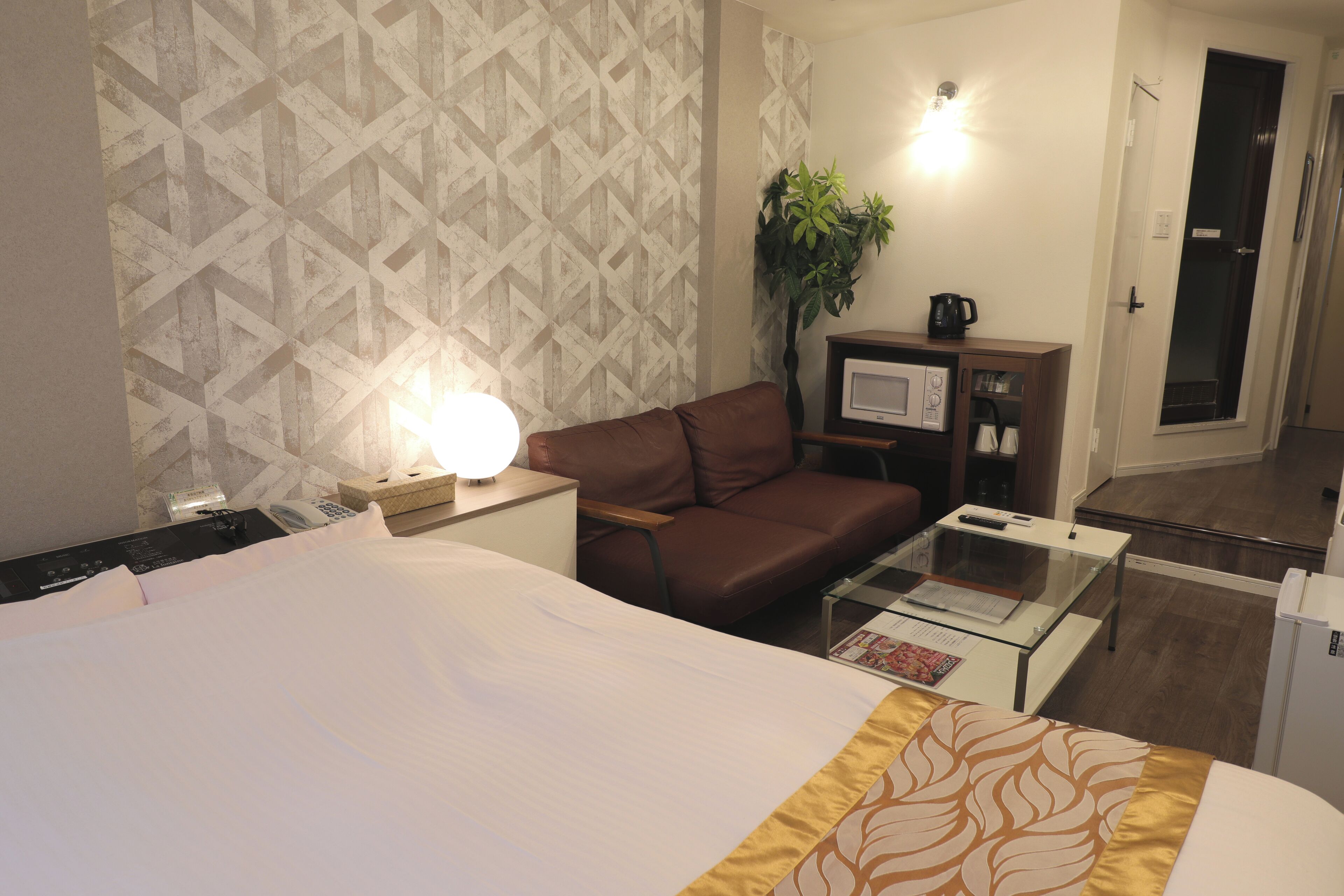 Habitación de hotel con cama, sofá de cuero marrón, mesita de centro de vidrio, televisor, microondas, planta y puerta.