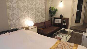 Habitación de hotel con cama, sofá de cuero marrón, mesita de centro de vidrio, televisor, microondas, planta y puerta.