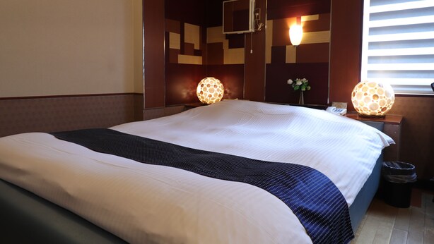 Een hotelkamer met een groot bed, twee bedlampen, een tafeltje met een telefoon en een raam met jaloezieën.