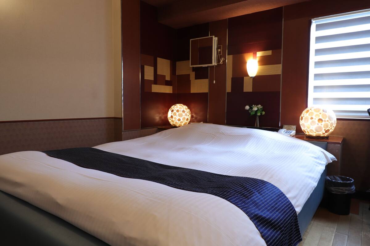 Une chambre d’hôtel avec un grand lit, deux lampes de chevet, une petite table avec un téléphone et une fenêtre avec des stores.