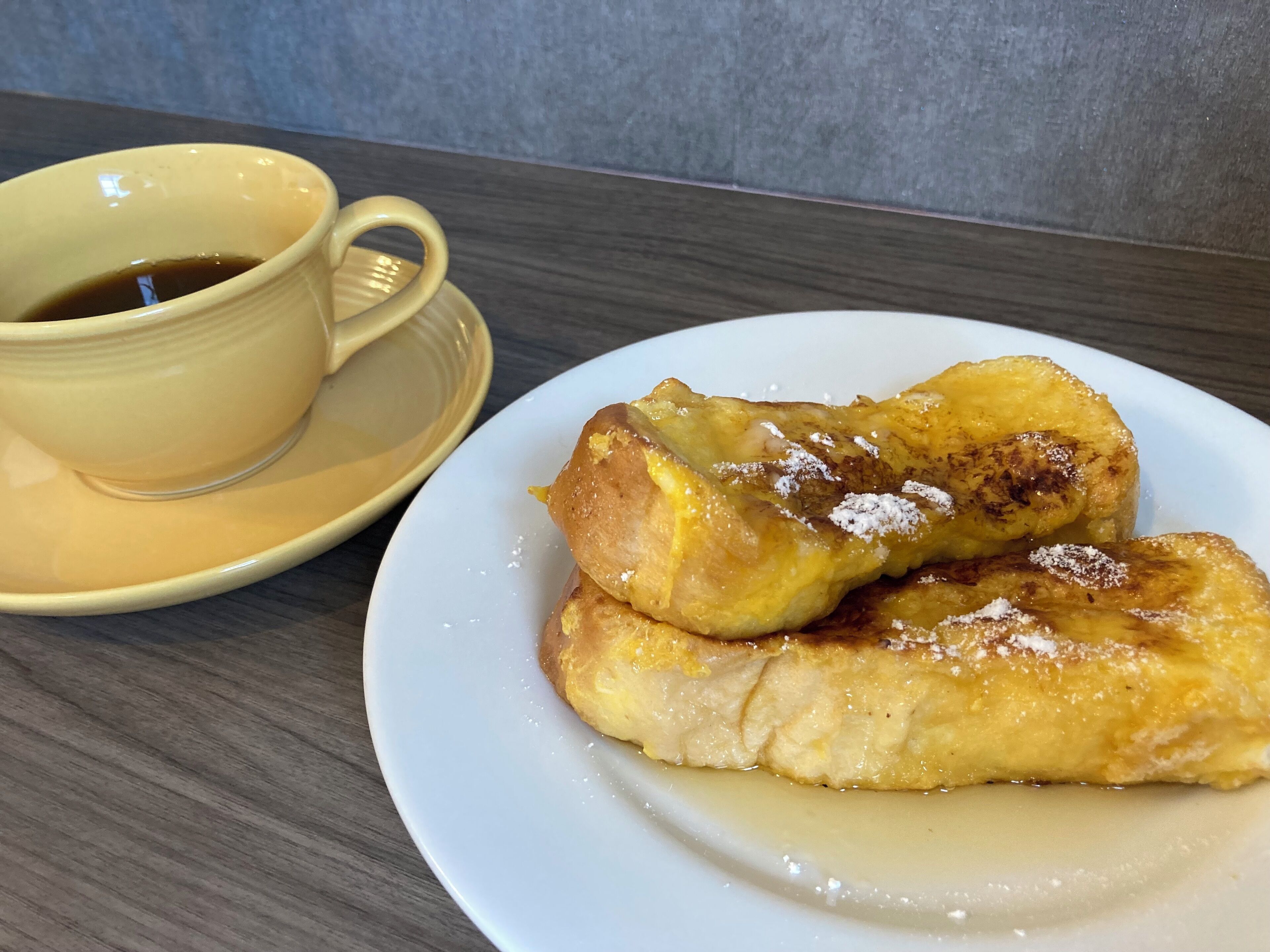 A plate of French toast with syrup and powdered sugar, and a cup of coffee on a saucer.