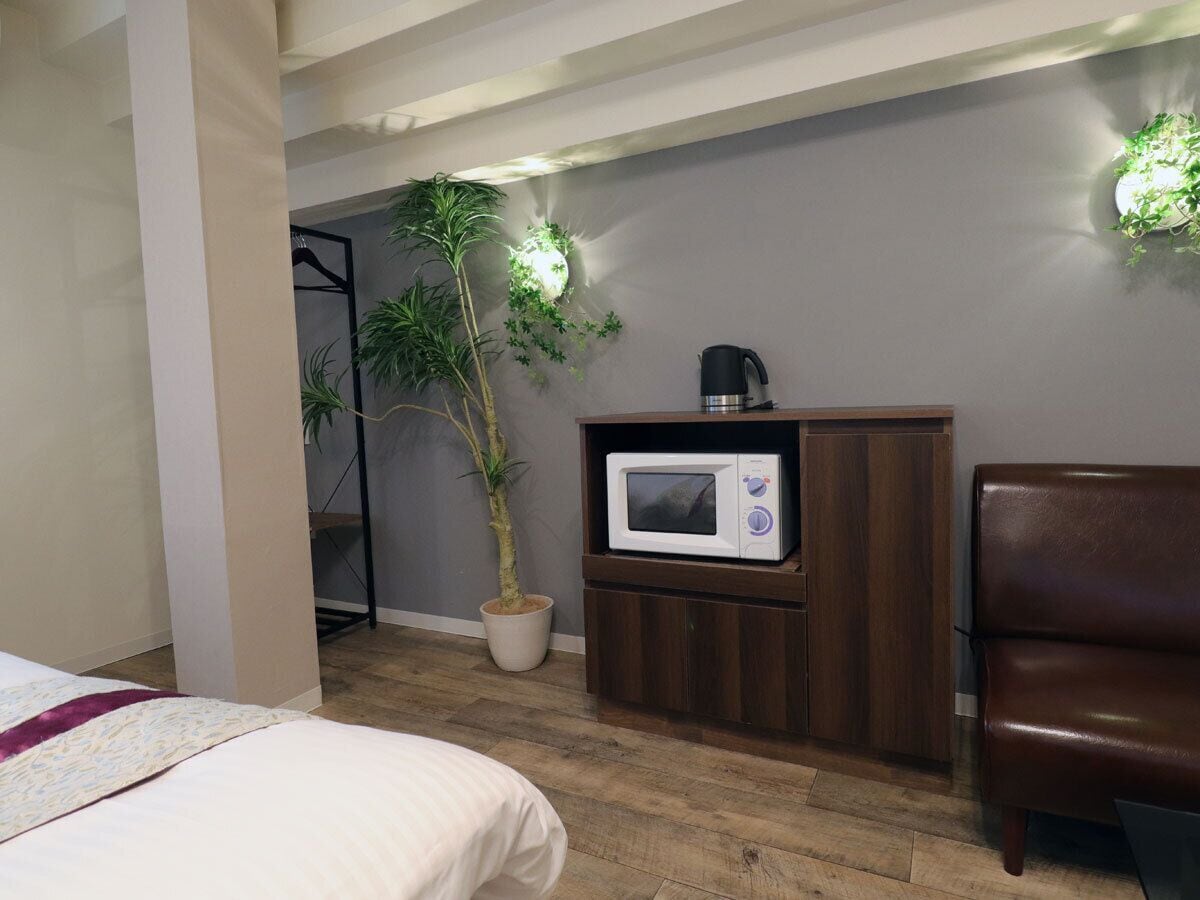 A hotel room with a bed, a microwave, a brown cabinet, a potted plant, and a chair.