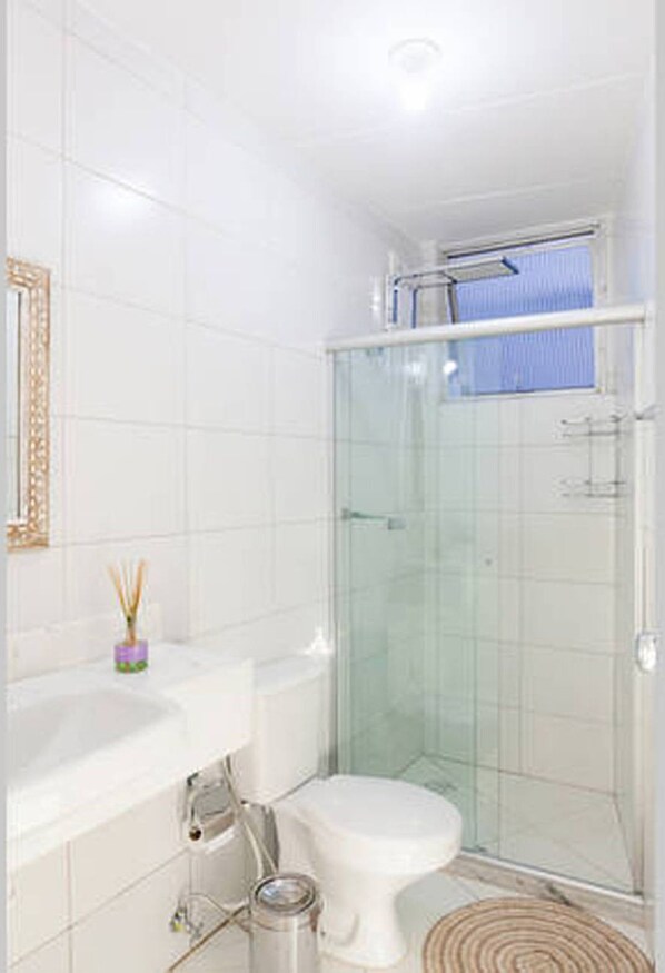 A small, modern bathroom with white tiles, a glass shower enclosure, a toilet, and a sink with a mirror above it. - Beautiful, modern and spacious, each room comes with a stunning sea view. (Salvador)