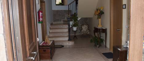 A hallway with a wooden door, a fire extinguisher, a staircase, a chandelier, and a vase with flowers.