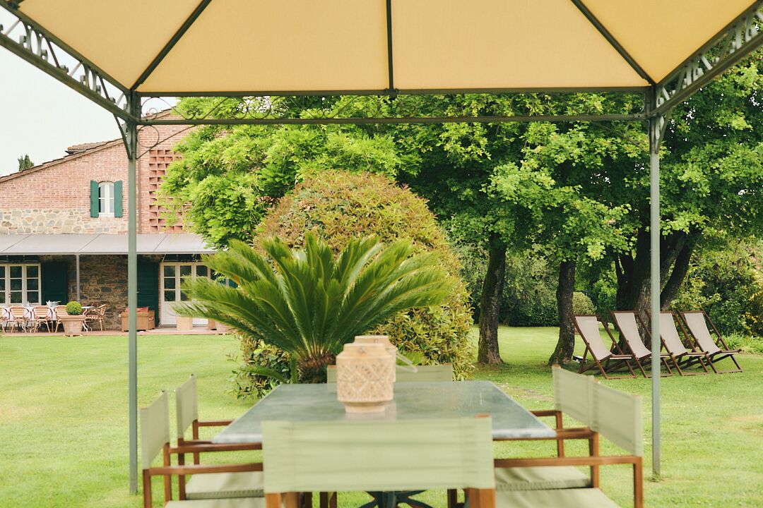 Un espace extérieur abrité, avec une table et des chaises, entouré de verdure et avec un bâtiment en arrière-plan.