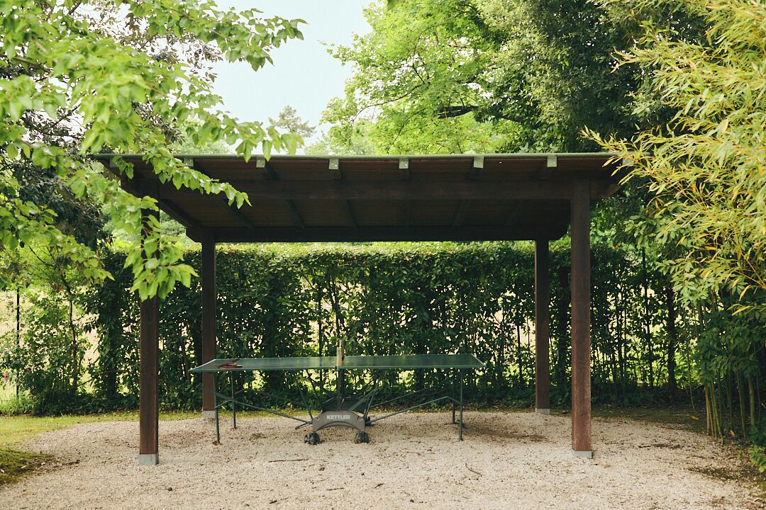 A covered outdoor seating area with a table and chairs, surrounded by trees.