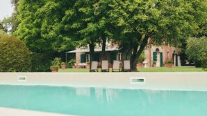 Ein Pool mit Liegestühlen, ein Backsteinhaus mit grünen Fensterläden und große Bäume im Hintergrund.