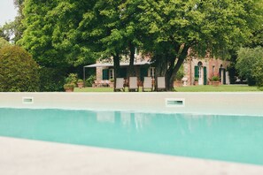 Ein Pool mit Liegestühlen, ein Backsteinhaus mit grünen Fensterläden und große Bäume im Hintergrund.