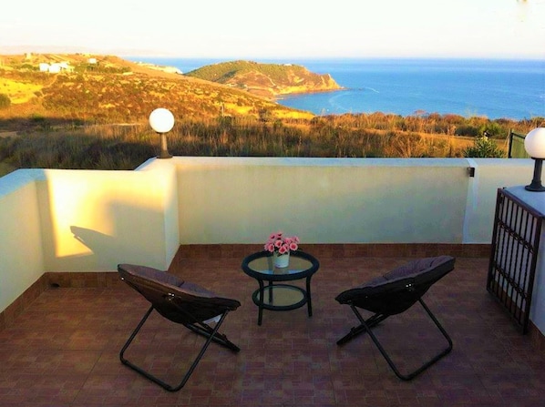 A balcony with two chairs and a small table, overlooking a coastal landscape with hills and the sea.