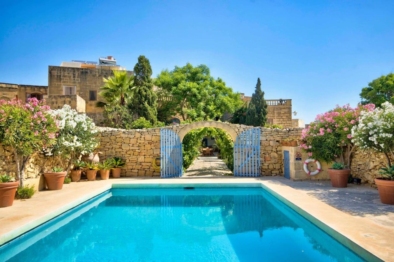 A swimming pool with a stone wall and archway entrance, surrounded by potted plants and trees.