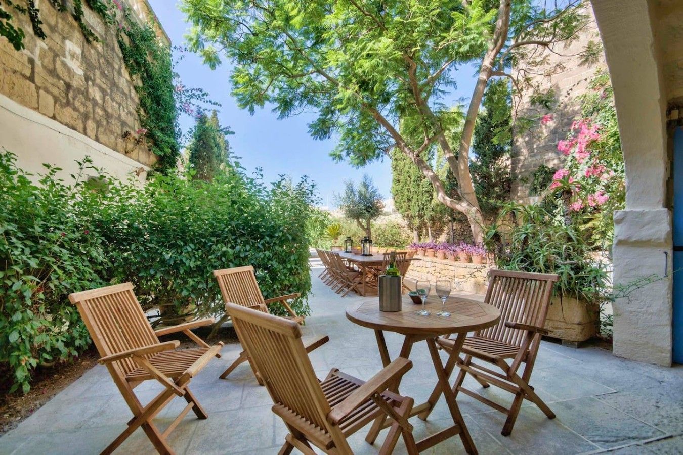 An outdoor seating area with wooden chairs and tables, surrounded by greenery and stone walls.