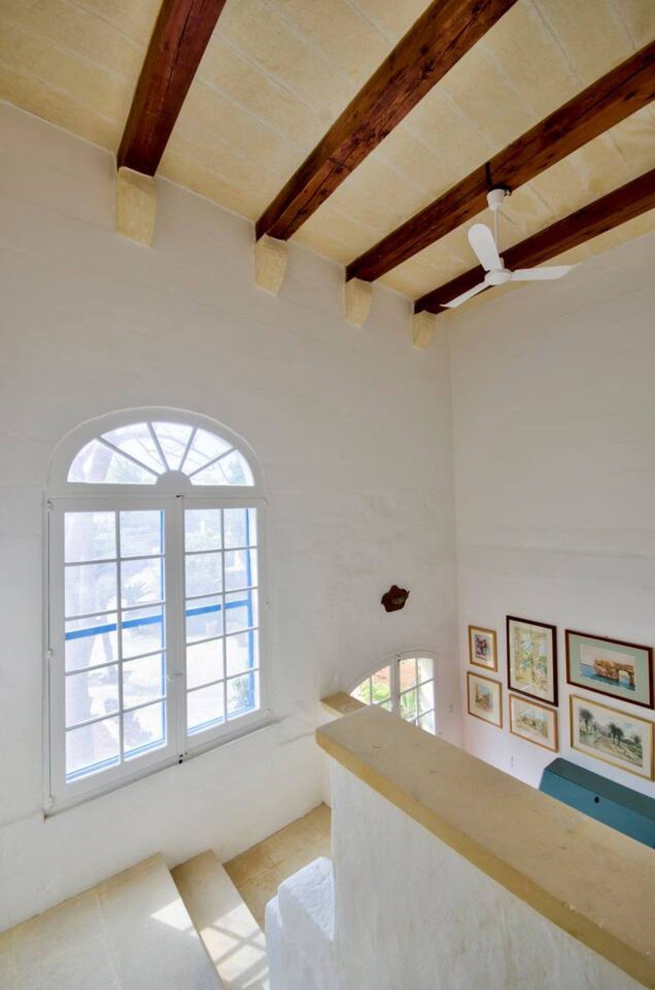 A room with a high ceiling, wooden beams, a large arched window, and a wall with framed pictures.