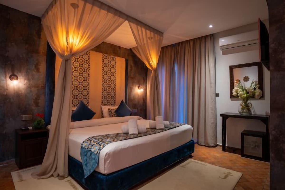 Un dormitorio con una cama grande, dosel, papel tapiz estampado, una mesita de noche, un jarrón con flores y un espejo en la pared.