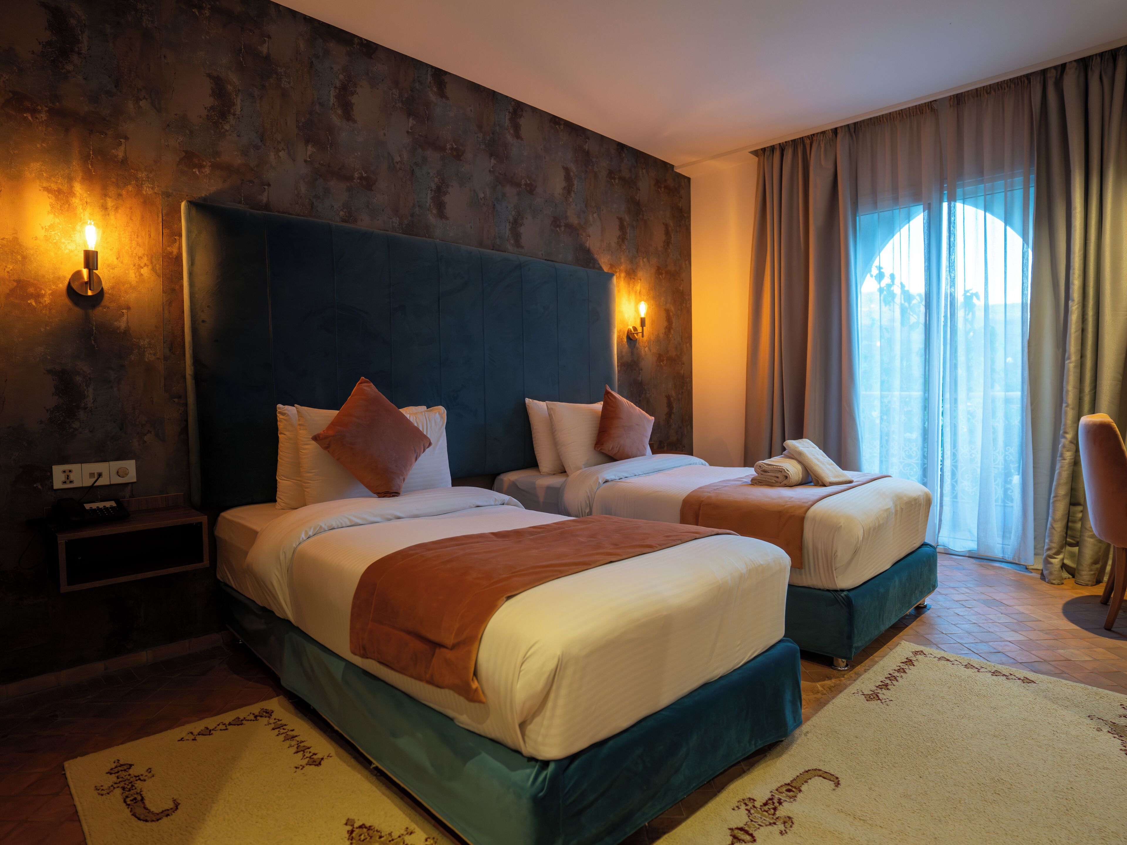 A hotel room with two beds, a large headboard, and a view of the outdoors.