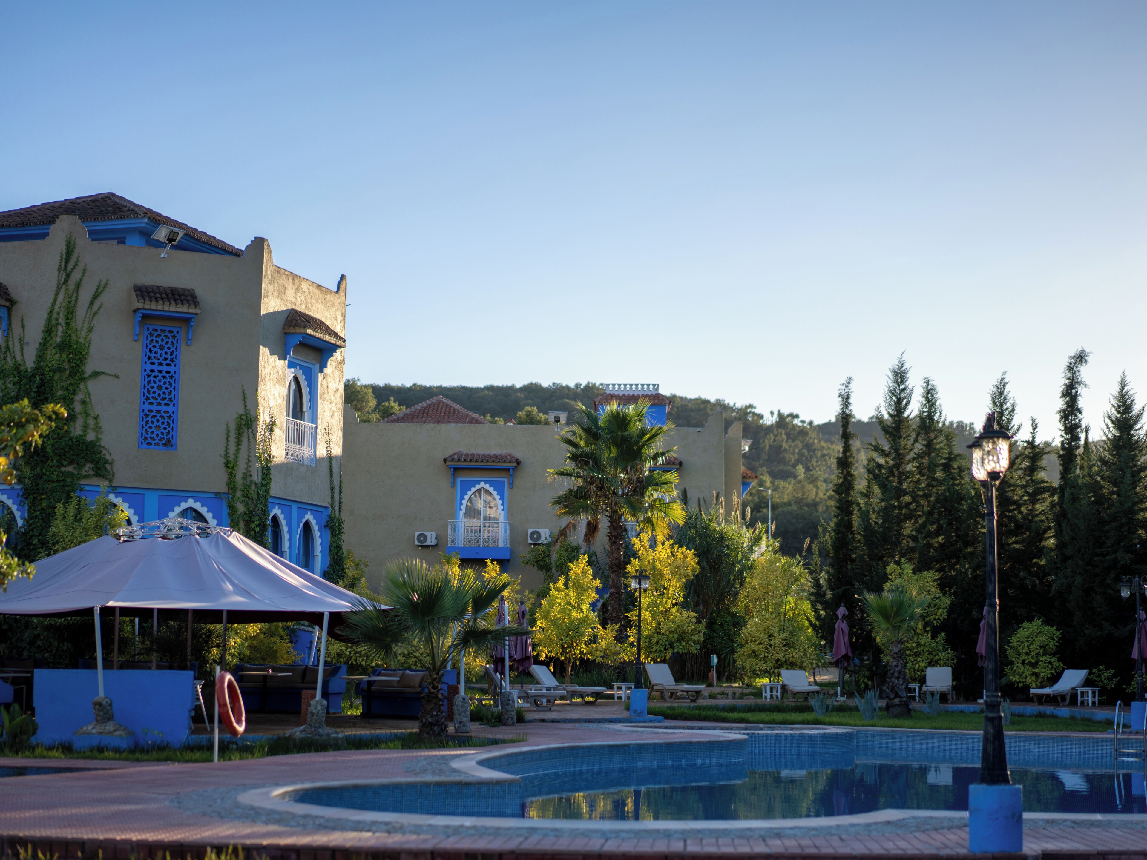 A pool area with lounge chairs, a covered seating area, and a building with blue accents and arched windows.